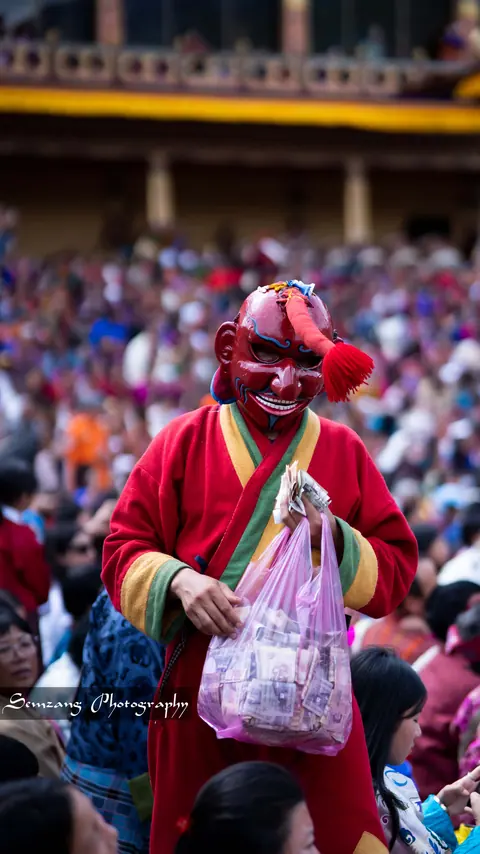Masked atsara clown in red robes carrying a plastic bag among the festival crowd