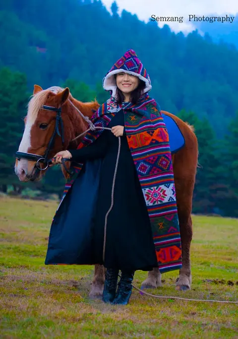 Woman in a patterned hooded cloak standing beside a horse in a mountain meadow