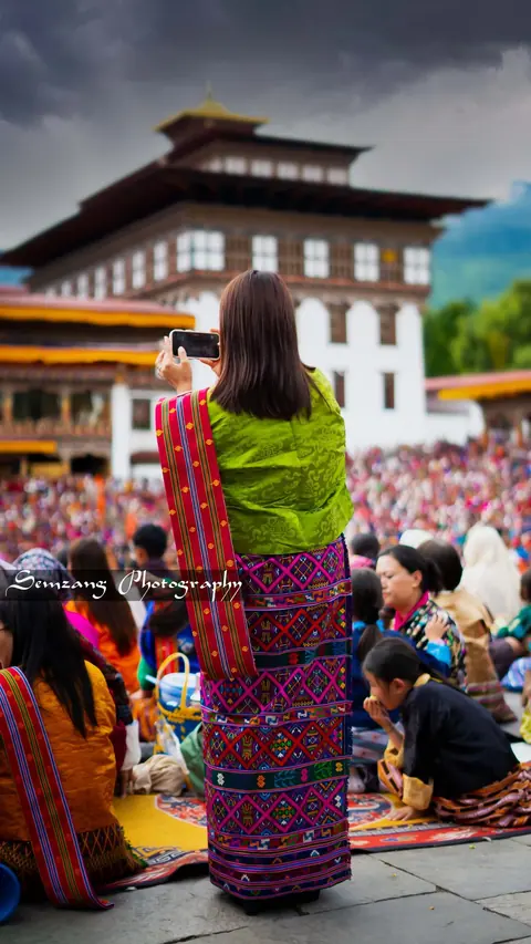 Woman in a green and pink kira seated at a festival in front of a dzong